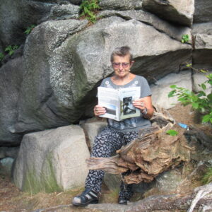 Starsza kobieta siedzi na kamieniu i czyta książkę, w tle skały