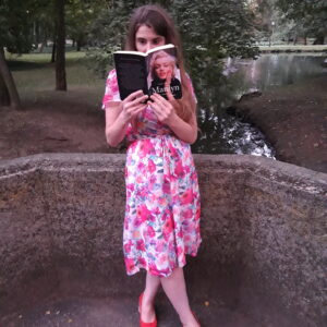 Młoda kobieta w kwiecistej sukience stoi i czyta książkę, w tle fragment parku