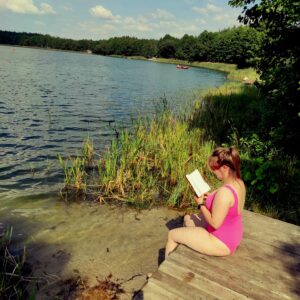 Młoda kobieta siedząca na pomoście czyta książkę, w tle jezioro.