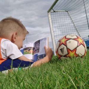 Chłopiec leży na murawie i czyta książkę, obok piłka