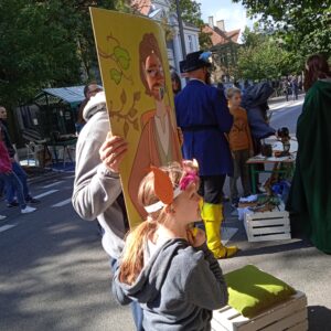 mężczyzna w okularach pokazuje twarz przez otwór w dużym obrazie przedstawiającym hobbita, obok stoi dziewczynka z dużymi uszami z papieru