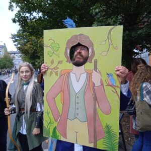 mężczyzna w okularach i z brodą pokazuje twarz przez otwór w dużym obrazie przedstawiającym hobbita, obok stoi kobiet z łukiem przebrana za elfa
