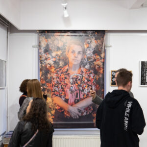 Na ścianie zawieszone duże zdjęcie portretowe kobiety. Po prawej i lewej stoją dwie osoby oglądające wystawę.