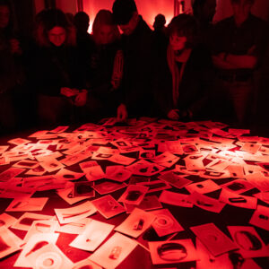 Zdjęcie z wernisażu wystawy Jarosława Klupsia. Oświetlenie czerwone, przypominające ciemnię fotograficzną. Stół. Na stole rozłożone zdjęcia różnych przedmiotów: żarówek, kuwet fotograficznych i innych. Przy stole stoi grupa osób.