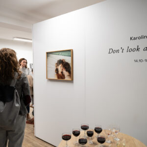 Zdjęcie w pomieszczeniu. Na pierwszym planie ściana z napisem: Don't look at me, Karolina Ćwik. Po lewej stronie widoczne zdjęcie zawieszone na ścianie w ramie oraz osoby zwiedzające wystawę.