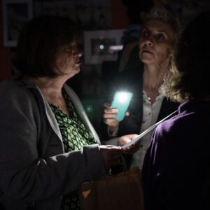 Dwie osoby stoją w ciemnym pomieszczeniu i oświetlają latarką z komórki kartkę papieru, na które jest coś napisane.