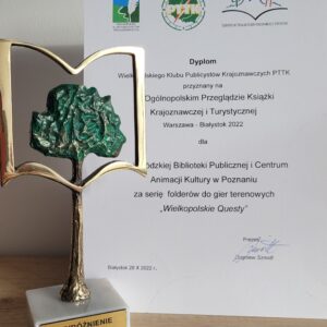 statuetka - drzewko w ramce w kształcie otwartej książki ustawiona na tle dyplomu dla WBPiCAK za serię folderów do gier terenowych 