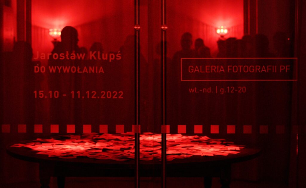 Zdjęcie przedstawia szklane drzwi wejściowe do galerii oświetlone czerwonym światłem. W drzwiach odbijają się stojące postacie.