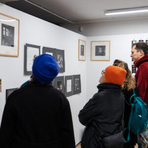 Pięć osób ustawionych tyłem lub lewych profilem ogląda wystawę czarno-białych fotografii, które wiszą na białych ściankach. Niektóre z nich oprawione są w jasne, drewniane ramy, część wisi bezpośrednio na ścianach.