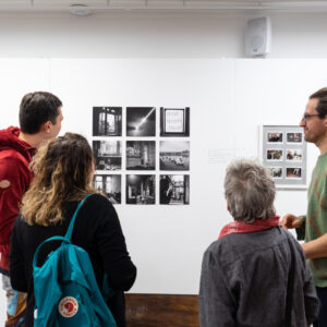 Zdjęcie przedstawia cztery osoby stojące przed białą ścianą, na której prezentowana jest wystawa fotografii. Trzy osoby stoją tyłem i patrzą na zdjęcia, a mężczyzna po prawej stoi bokiem i patrzy w lewo w stronę tych osób.