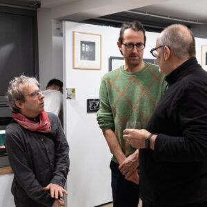 Trzech dorosłych mężczyzn rozmawia ze sobą, stojąc w przestrzeni wystawy fotografii. W tle widać dwie inne osoby.