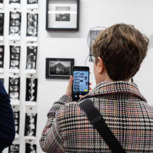 Zdjęcie przedstawia kobietę w krótkich włosach i płaszczu, stojącą tyłem i robiącą telefonem zdjęcie wystawy fotografii, które znajdują się przed nią. Po lewej stronie widać fragment innej osoby.