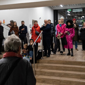 Zdjęcie przedstawia grupę ponad dwudziestu osób, która stoi w przestronnym holu biblioteki, przed niewysokimi schodami. Wszyscy skierowani są w stronę stojącego tyłem mężczyzny, znajdującego się w lewej dolnej części zdjęcia