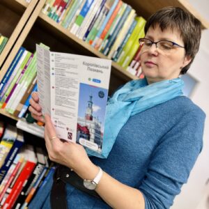 Na zdjęciu kobieta w niebieskiej sukni czyta ulotkę Wielkopolskich Questów na tle książek w czytelni.