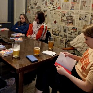 grupa osób siedzi przy długim stole, na którym są książki, notesy i napoje, jedna kobieta trzyma otwartą książkę