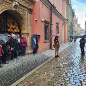 kamienica z ozdobnym portalem przy brukowanej ulicy, obok wejścia stoją ludzie, kilka osób trzyma otwarte parasole