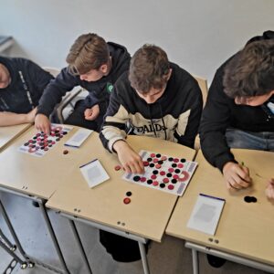 czworo chłopców siedzi przy stolikach, na których są rozłożone plansze do gry i kolorowe żetony