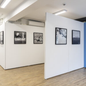 Widok wystawy - na ścianach wiszą czarno-białe fotografie w czarnych ramach, po prawej stronie na niebieskiej ściance znajduje się tytuł wystawy wraz z opisem.