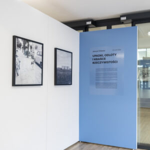Widok wystawy - na białej ścianie po lewej wiszą dwie czarno-białe fotografie w czarnych ramach, po prawej stronie na niebieskiej ściance znajduje się tytuł wystawy wraz z opisem.